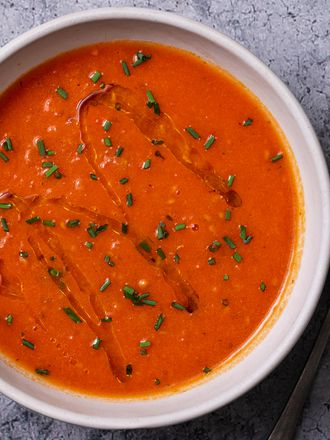 A bowl of creamy vegan tomato soup on a stone background with a spoon to the right of the bowl. The surface of the soup is drizzled with olive oil and sprinkled with chopped herbs.